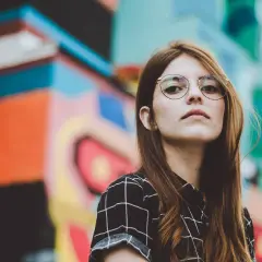 Młoda kobieta w okularach dla krótkowidza, z rudymi włosami, na tle kolorowego graffiti.