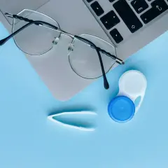 Okulary, pojemnik na soczewki i pęseta na tle laptopa. Czy można nosić soczewki i okulary jednocześnie? To zależy od wady wzroku.