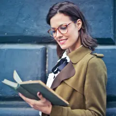 Kobieta w okularach z lekką wadą wzroku 0.25 czyta książkę, opierając się o niebieską ścianę.