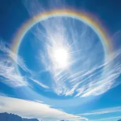 Słońce otoczone tęczowym halo na tle błękitnego nieba z chmurami. Jak usunąć efekt halo? To zjawisko atmosferyczne, nie da się go usunąć.