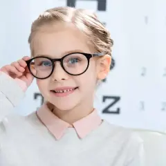 Uśmiechnięta dziewczynka w okularach przed tablicą optometryczną. Czy wzrok może się poprawić? Tak, dzięki odpowiedniej korekcji!