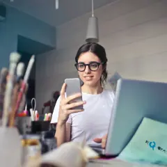Kobieta w okularach do laptopa przegląda coś na telefonie, siedząc przy biurku z laptopem i pędzlami.