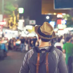 Mężczyzna w kapeluszu i z plecakiem idzie nocą przez tętniący życiem bazar. To jego widzenie w ciemności, pełne kolorowych świateł i ludzi.
