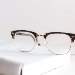 Okulary z oprawkami w stylu retro, idealne do korygowania wady wzroku +2. Leżą na białej książce.