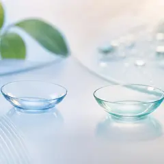 Dwie soczewki silikonowo-hydrożelowe na jasnym tle z kroplami wody i zielonym liściem.