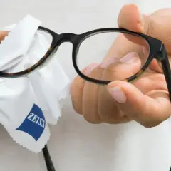 Dłonie przecierają okulary białą ściereczką z logo Zeiss, by zapewnić czysty obraz.