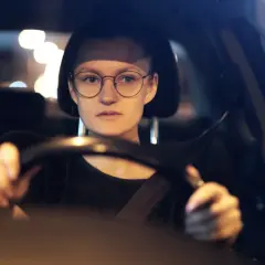 Kobieta w okularach prowadzi samochód nocą. Rozmazane światła uliczne sugerują, że może mieć kurzą ślepotę, ale okulary pomagają jej widzieć.
