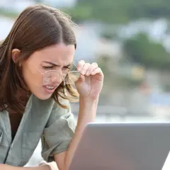Kobieta z wadą wzroku -0.75, z trudem czyta tekst na laptopie, poprawiając okulary.