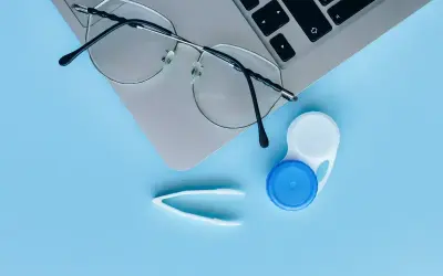 Okulary, pojemnik na soczewki i pęseta na tle laptopa. Czy można nosić soczewki i okulary jednocześnie? To zależy od wady wzroku.