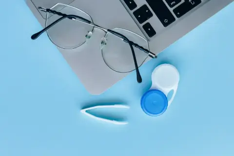 Okulary, pojemnik na soczewki i pęseta na tle laptopa. Czy można nosić soczewki i okulary jednocześnie? To zależy od wady wzroku.