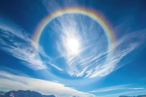 Słońce otoczone tęczowym halo na tle błękitnego nieba z chmurami. Jak usunąć efekt halo? To zjawisko atmosferyczne, nie da się go usunąć.
