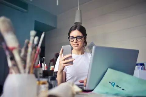 Kobieta w okularach do laptopa przegląda coś na telefonie, siedząc przy biurku z laptopem i pędzlami.
