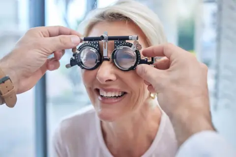 Kobieta uśmiecha się podczas badania wzroku, gdzie optometrysta dopasowuje cieniowanie szkieł w okularach próbnych.