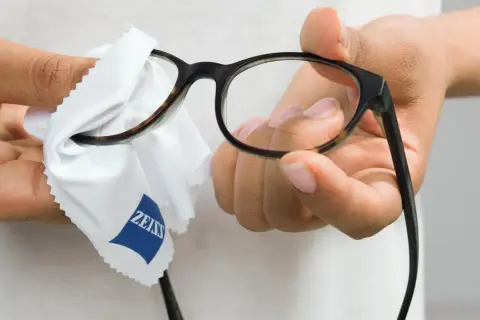 Dłonie przecierają okulary białą ściereczką z logo Zeiss, by zapewnić czysty obraz.