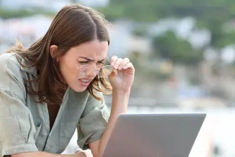 Kobieta z wadą wzroku -0.75, z trudem czyta tekst na laptopie, poprawiając okulary.