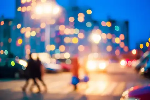 Rozmyte światła miasta nocą, jak widzi osoba z wadą wzroku. Dwie sylwetki przechodzą przez ulicę, otoczone kolorowymi plamami.