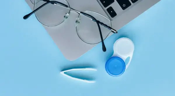 Okulary, pojemnik na soczewki i pęseta na tle laptopa. Czy można nosić soczewki i okulary jednocześnie? To zależy od wady wzroku.