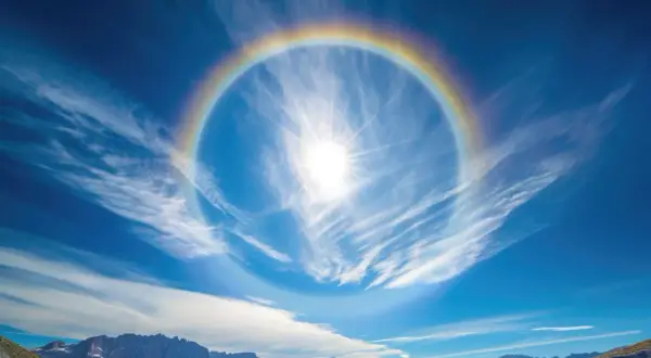 Słońce otoczone tęczowym halo na tle błękitnego nieba z chmurami. Jak usunąć efekt halo? To zjawisko atmosferyczne, nie da się go usunąć.