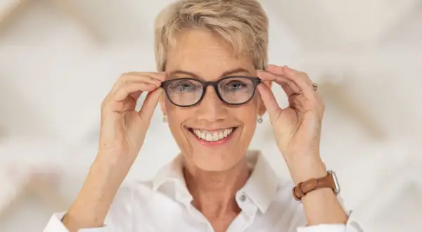 Uśmiechnięta kobieta z krótkimi blond włosami poprawia okulary na nosie. Okulary na zeza pomagają jej widzieć świat wyraźniej.
