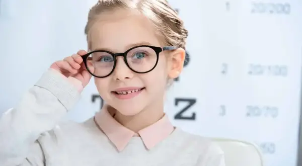 Uśmiechnięta dziewczynka w okularach przed tablicą optometryczną. Czy wzrok może się poprawić? Tak, dzięki odpowiedniej korekcji!