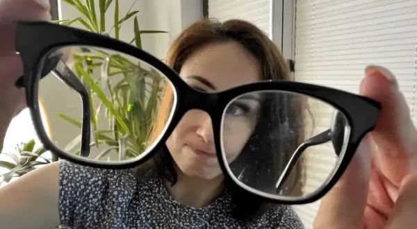 Kobieta trzyma okulary, zastanawiając się, czy można myć okulary płynem do naczyń. W tle roślina i okno.