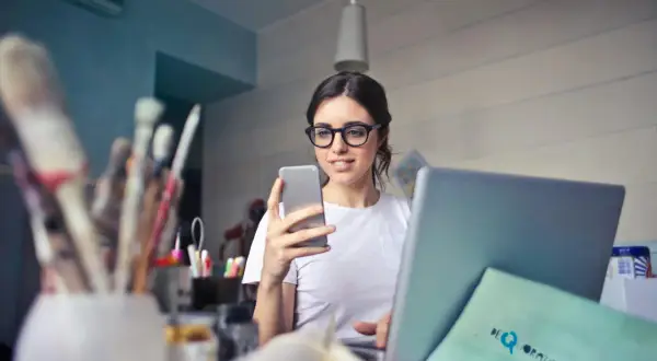 Kobieta w okularach do laptopa przegląda coś na telefonie, siedząc przy biurku z laptopem i pędzlami.