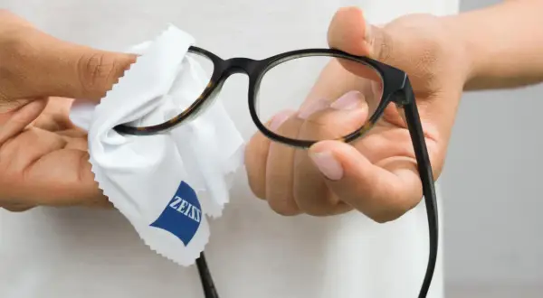 Dłonie przecierają okulary białą ściereczką z logo Zeiss, by zapewnić czysty obraz.