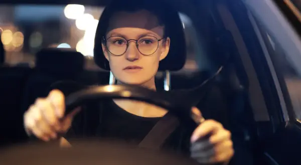 Kobieta w okularach prowadzi samochód nocą. Rozmazane światła uliczne sugerują, że może mieć kurzą ślepotę, ale okulary pomagają jej widzieć.