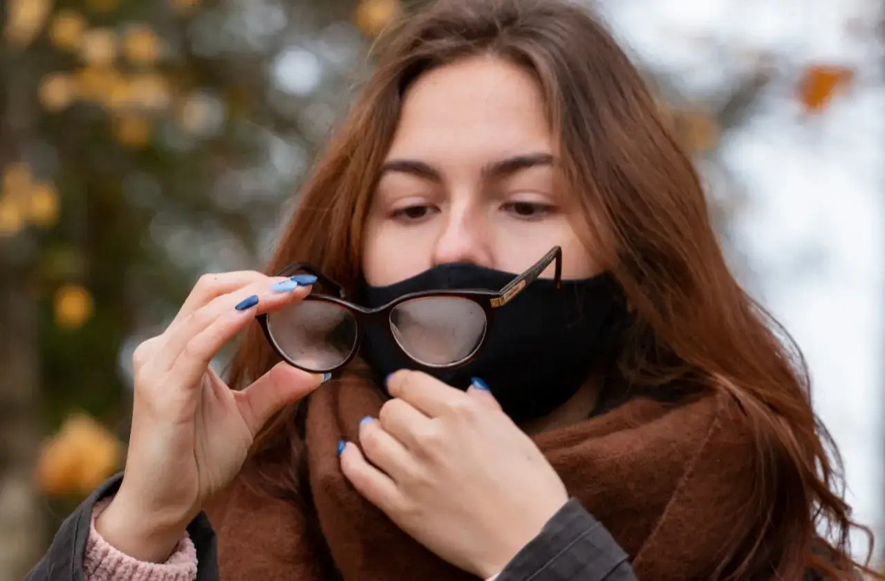 Dziewczyna w masce i szaliku patrzy na zaparowane okulary, które trzyma w dłoni.