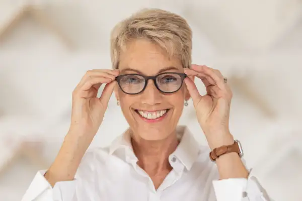 Uśmiechnięta kobieta z krótkimi blond włosami poprawia okulary na nosie. Okulary na zeza pomagają jej widzieć świat wyraźniej.