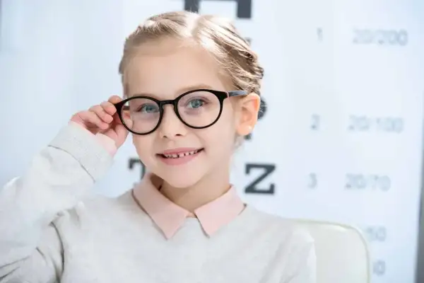 Uśmiechnięta dziewczynka w okularach przed tablicą optometryczną. Czy wzrok może się poprawić? Tak, dzięki odpowiedniej korekcji!