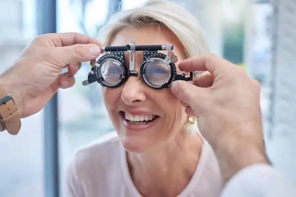 Kobieta uśmiecha się podczas badania wzroku, gdzie optometrysta dopasowuje cieniowanie szkieł w okularach próbnych.