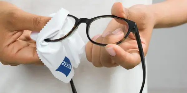 Dłonie przecierają okulary białą ściereczką z logo Zeiss, by zapewnić czysty obraz.