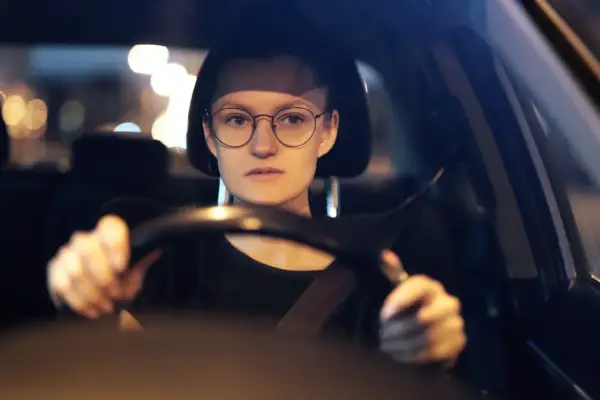 Kobieta w okularach prowadzi samochód nocą. Rozmazane światła uliczne sugerują, że może mieć kurzą ślepotę, ale okulary pomagają jej widzieć.